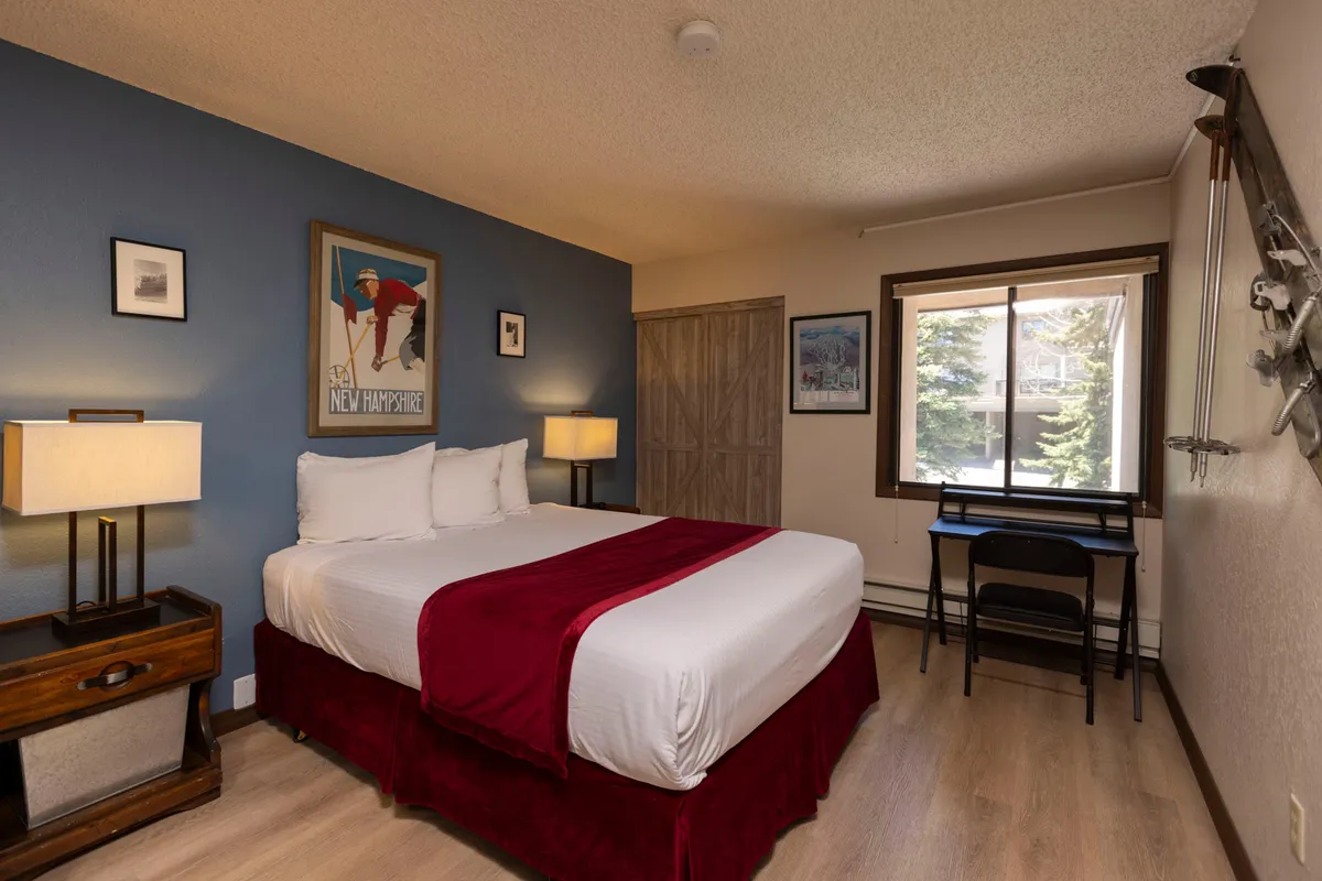 This image features a cozy queen bedroom with a warm red accent on the bed, framed artwork above the headboard, a desk and chair perfect for unwinding, and a sunlit window offering a tranquil view. The soft lighting and inviting textures evoke a sense of rest and relaxation, ideal for a restful night's sleep.