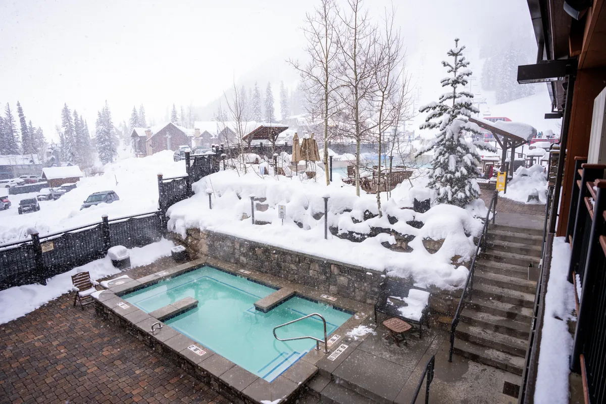 A tranquil outdoor hot tub surrounded by snow-covered landscape and evergreens, with steam rising against the wintry backdrop. People are seen relaxing and socializing, emphasizing a warm, inviting return after a day out.
