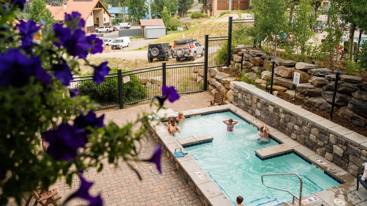 A lively exterior scene featuring a sparkling swimming pool surrounded by blooming flowers and people enjoying the water. The atmosphere is cheerful and relaxed, suggesting a communal space to unwind after outdoor activities.