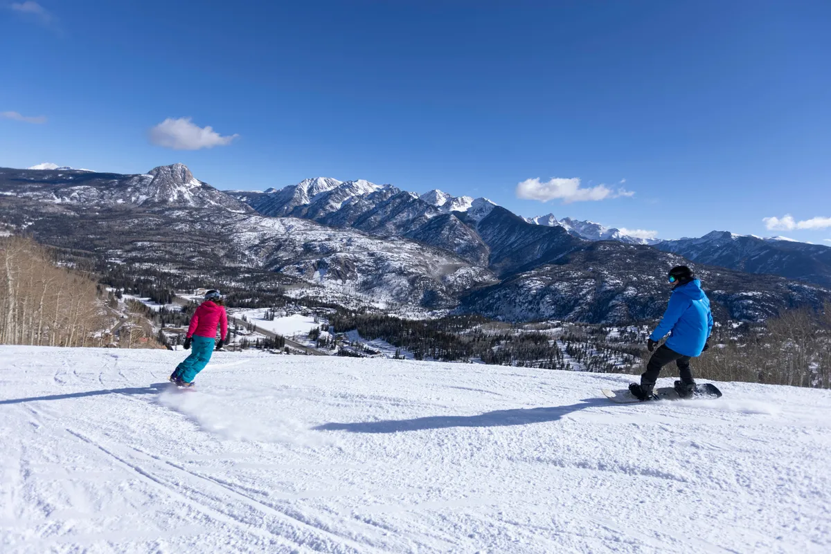 An exhilarating view of snowboarders descending the mountain against a backdrop of snowy peaks and a clear blue sky. The image radiates the thrill and beauty of a day spent on the slopes, inviting viewers to imagine themselves in the heart of the action.