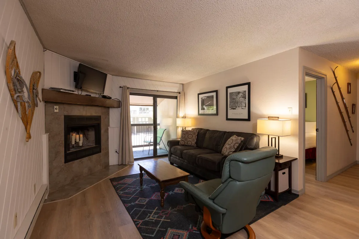 The image shows a warm, inviting living area featuring a cozy sofa set around a crackling fireplace. Sunlight pours in through a sliding glass door, illuminating a welcoming coffee table that's perfect for family games or morning coffee. The space is designed for relaxation and togetherness, with a comfortable, homey atmosphere.
