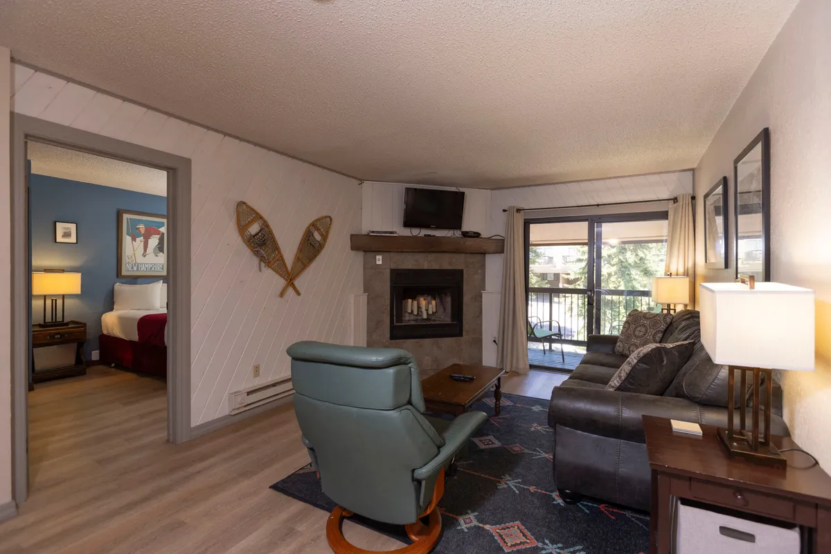 A cozy living room with a fireplace as its centerpiece, surrounded by comfortable seating. Large windows offer a glimpse of the outdoors, while the arrangement invites relaxation and social gatherings. The sofa and armchairs are positioned for easy conversation, and the overall atmosphere is warm and inviting.