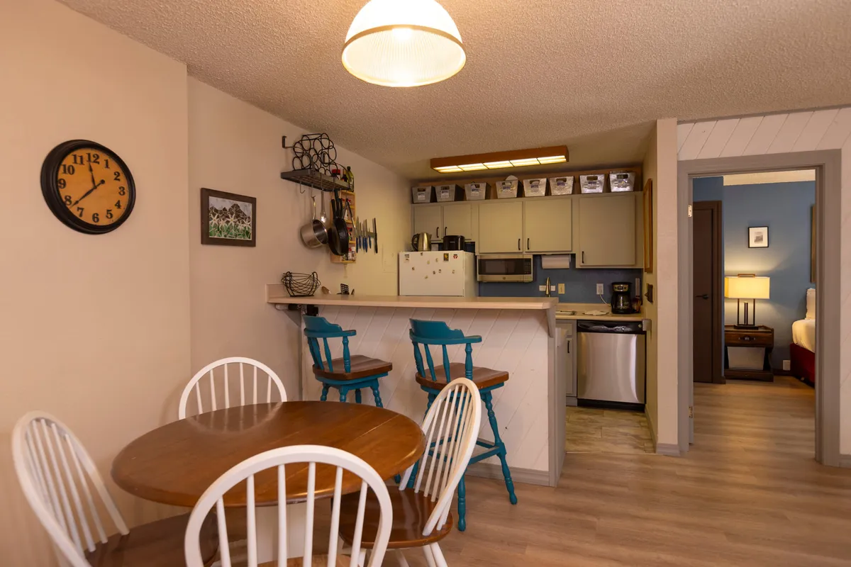 A bright and cheerful dining area featuring a dining table, bar stools at a kitchen counter, and a decorative clock on the wall. The open layout visually connects the dining space to the kitchen, reinforcing a sense of togetherness. The area is set up for family meals, casual gatherings, and entertaining.