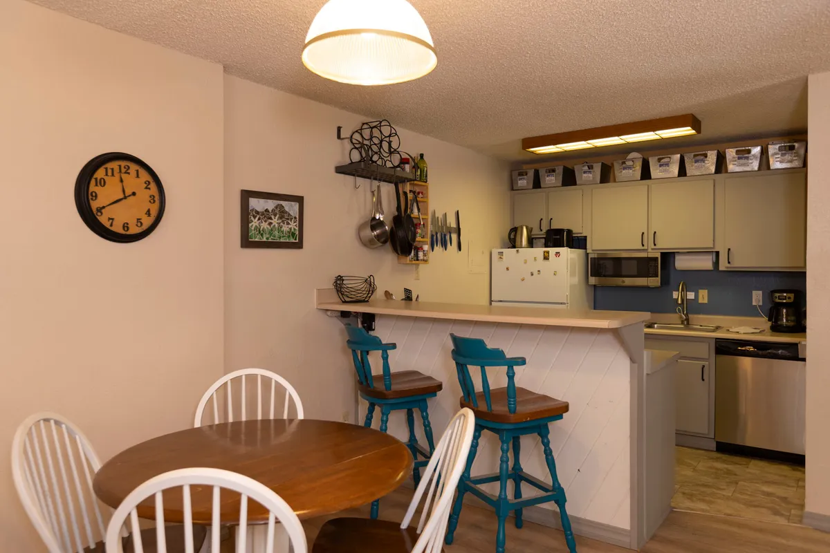 A cozy kitchen and dining nook with a wooden table and blue bar stools. The kitchen area is visible, suggesting a space where cooking and socializing naturally blend. The setup encourages both shared meals and casual interaction.