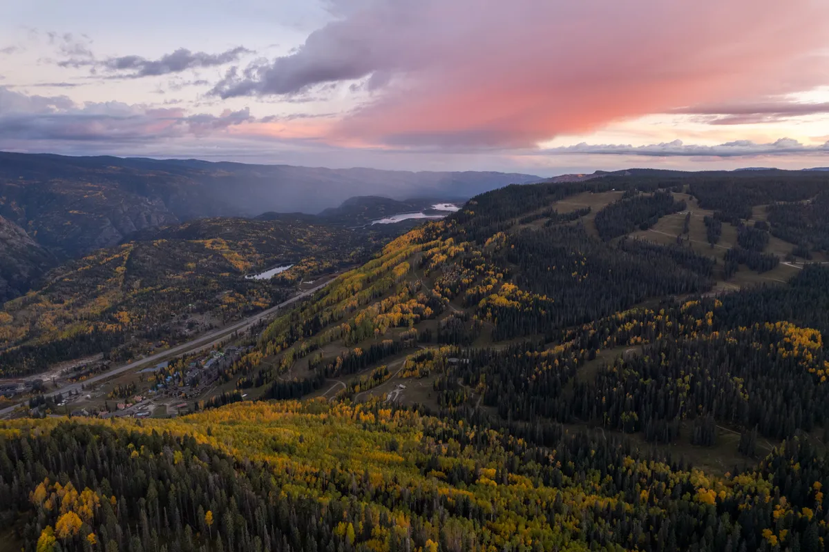 This image captures expansive rolling hills adorned with vibrant autumn foliage, distant mountain peaks, and a serene sunset sky. The landscape is open and inviting, suggesting a perfect environment for outdoor activities such as hiking and mountain biking.