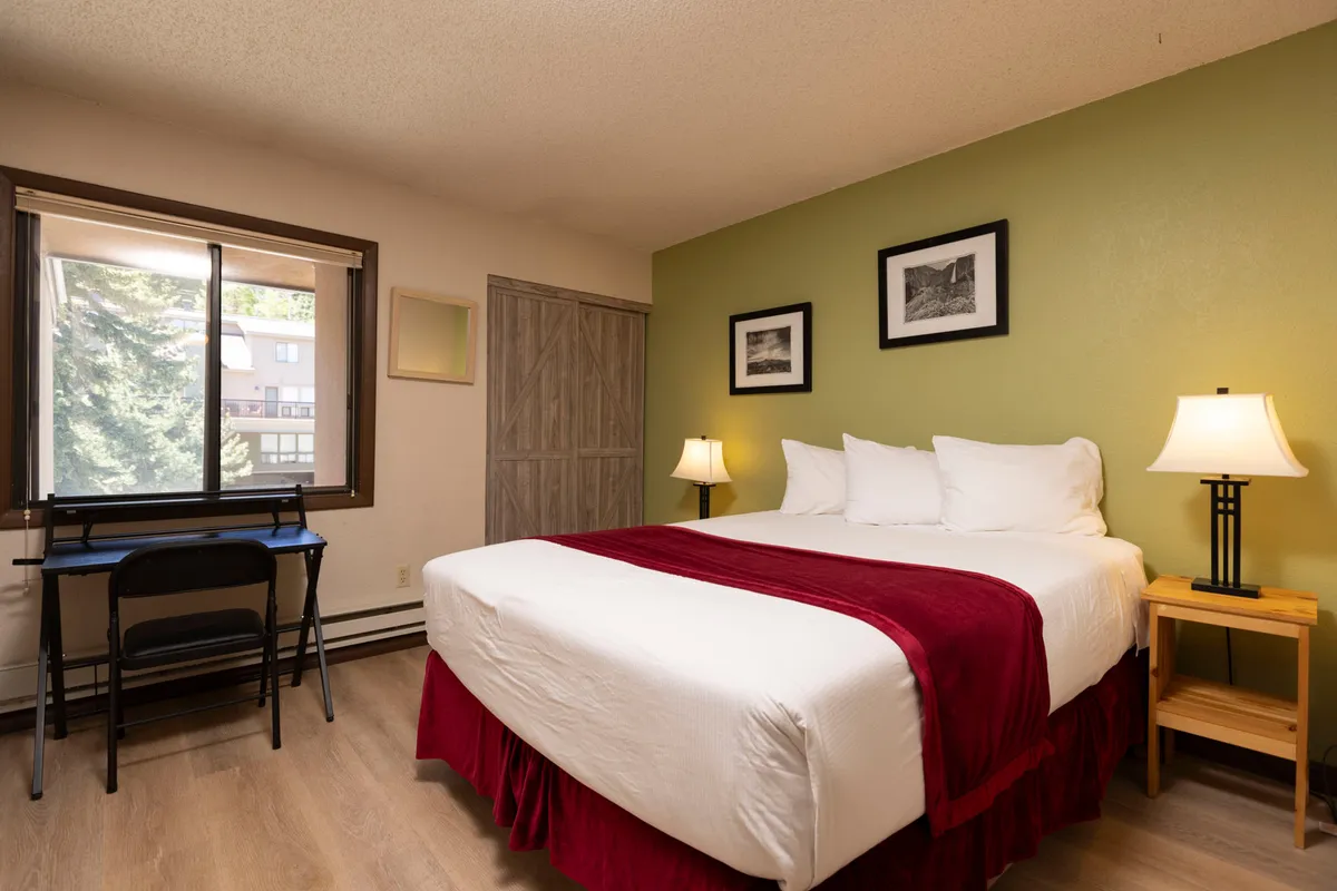 A second queen bedroom is depicted, featuring a plush bed with a red accent throw, bedside tables with lamps for reading, a desk and chair, and framed pictures that add a homely touch. The warm, cozy ambience encourages relaxation and comfort.